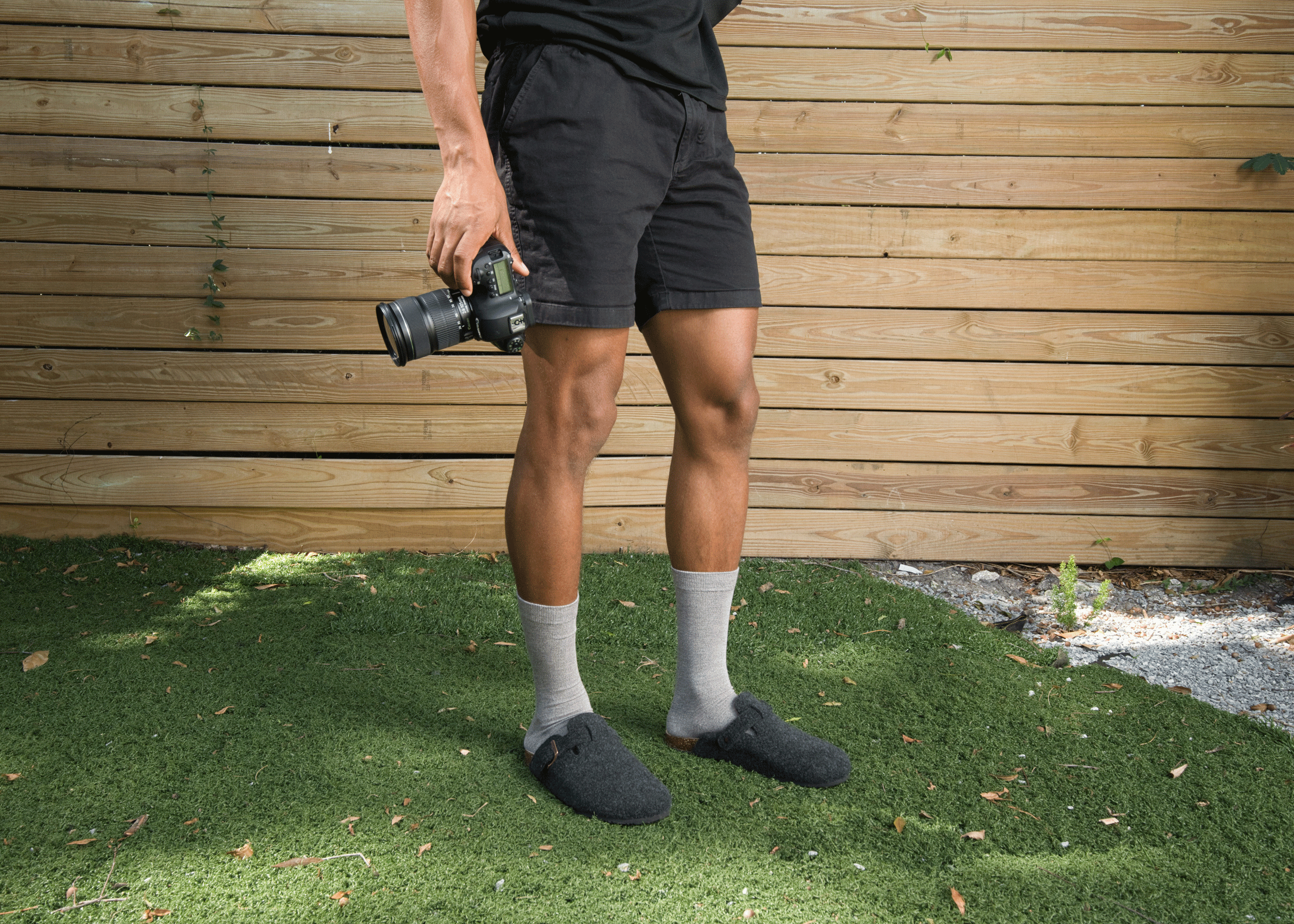 Person wearing black shorts and gray bamboo socks standing on grass with a wooden wall in the background