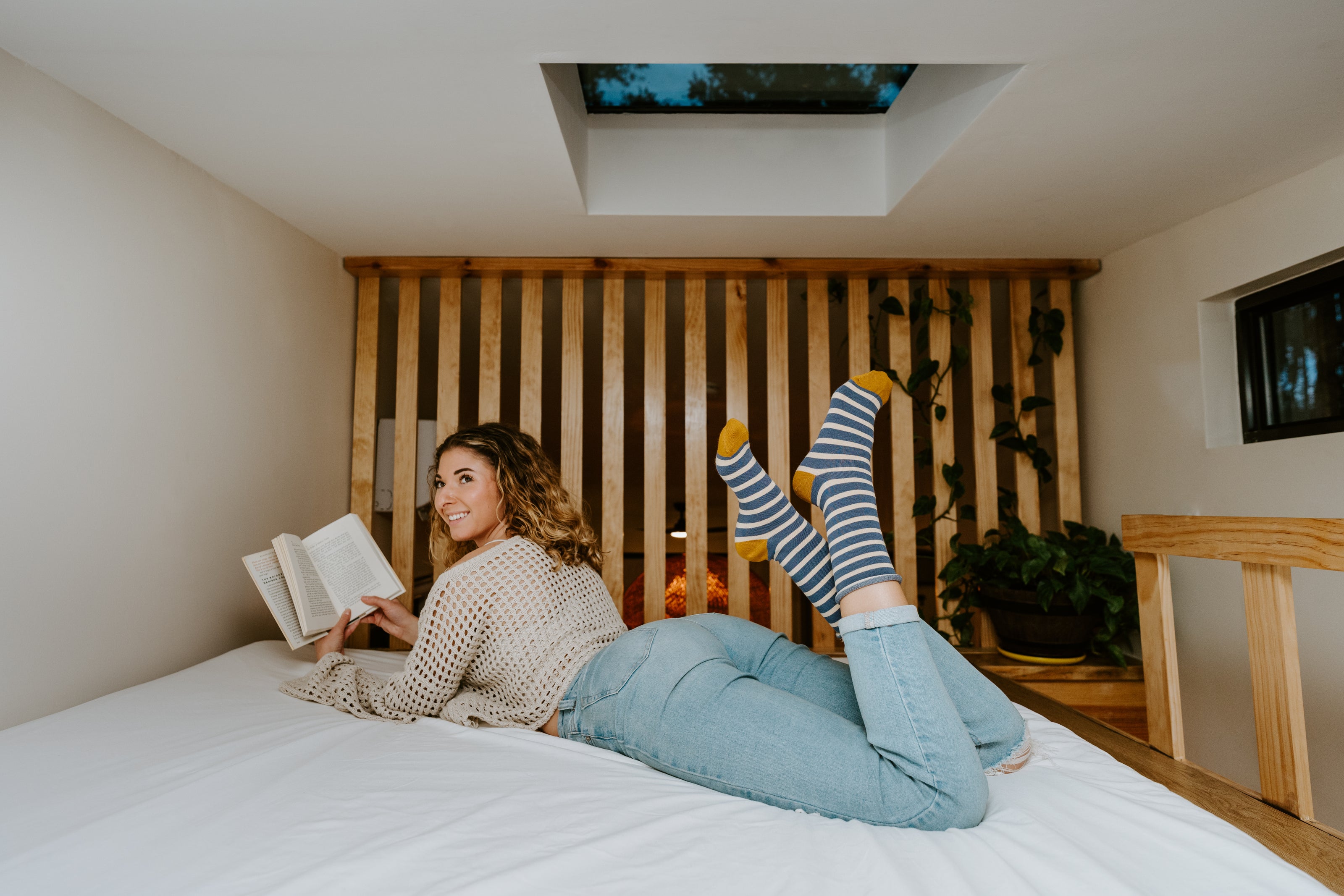 Woman lying on a bed reading a book in a cozy room with wooden headboard and plants wearing blue and gold striped bamboo socks.