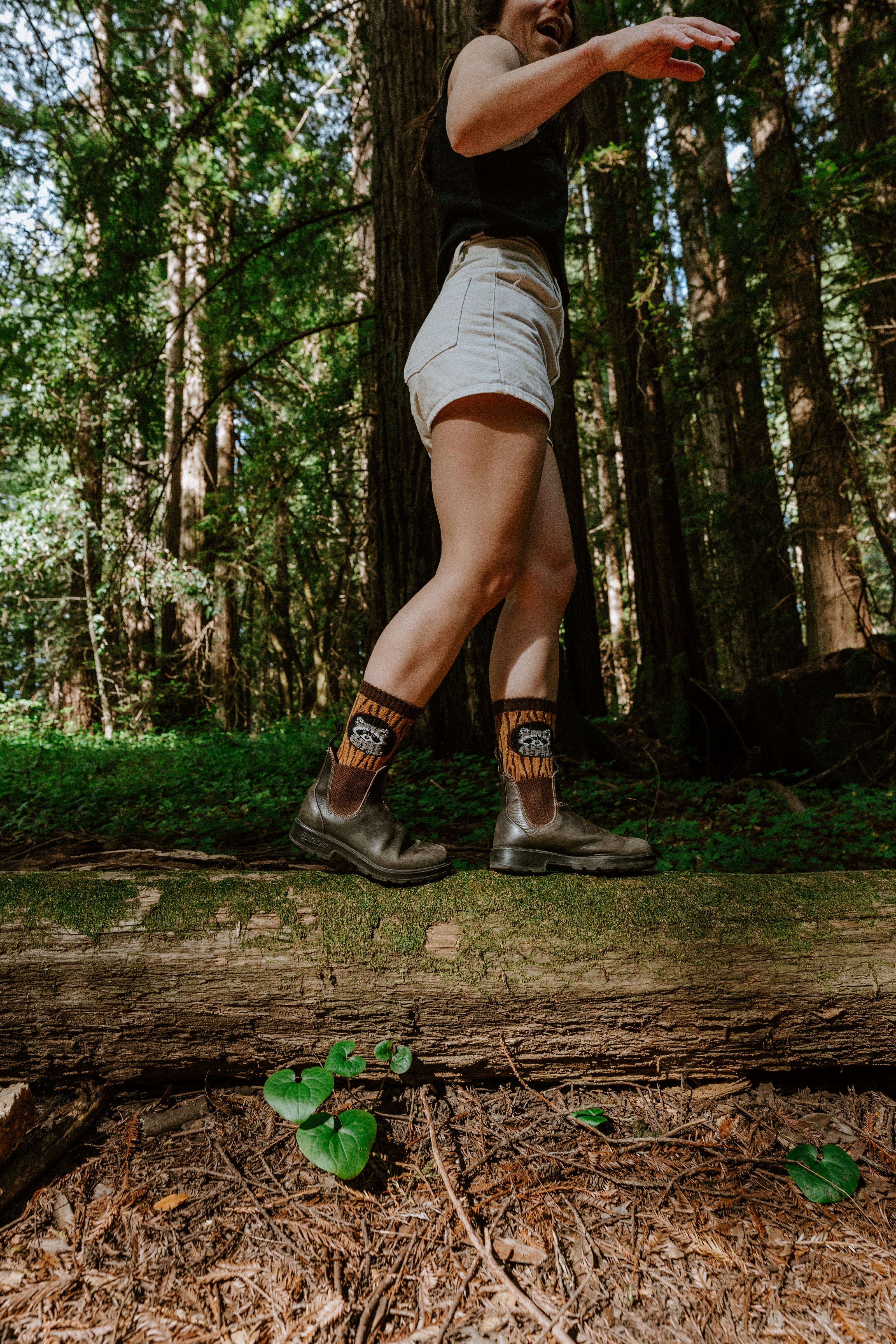 A person walking on a log in a forest, wearing brown boots, light denim shorts, and patterned socks with a raccoon graphic.