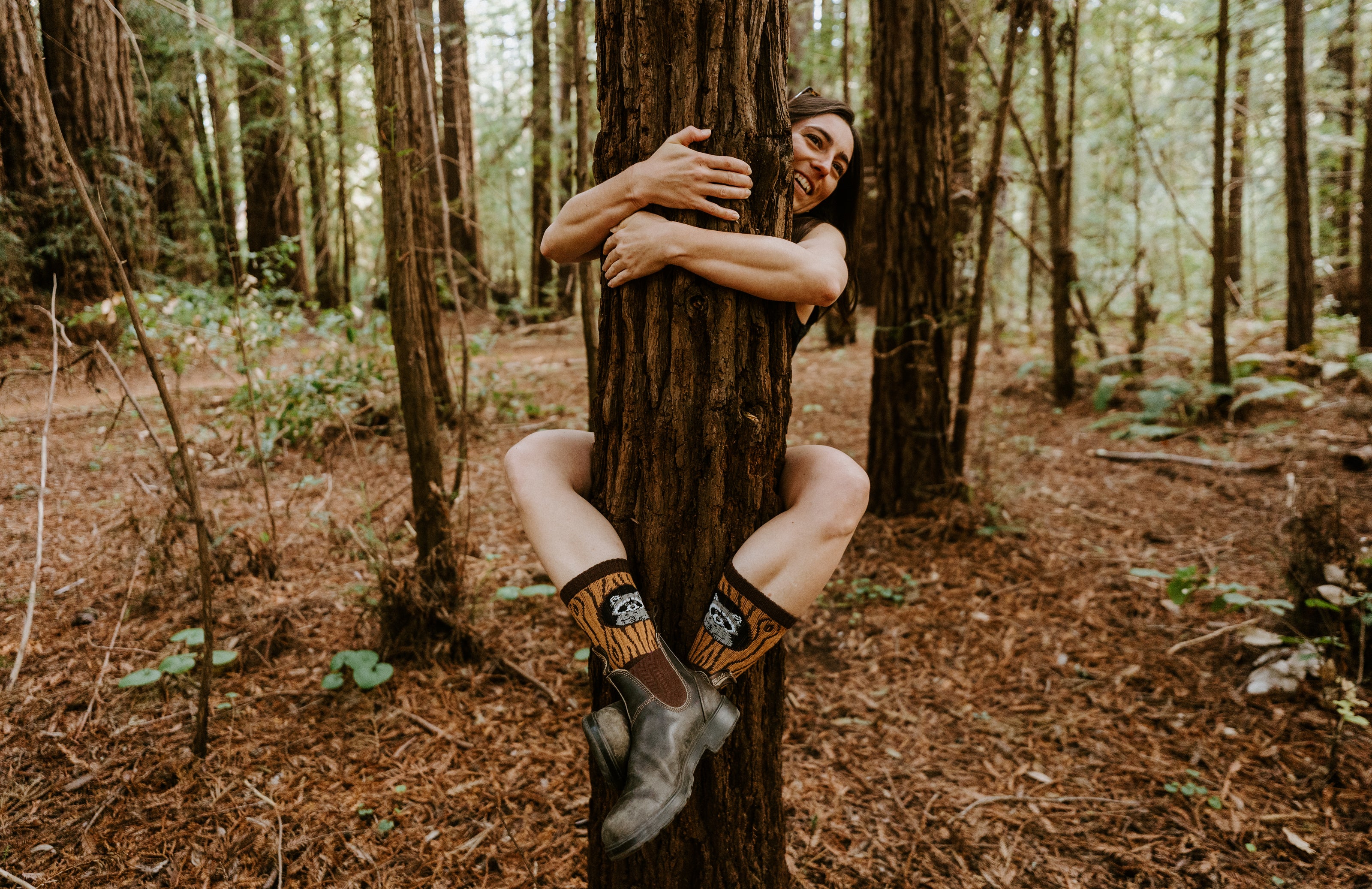 A woman in a forest setting, hugging a large tree trunk wearing raccoon-themed Trash Bandit socks. 