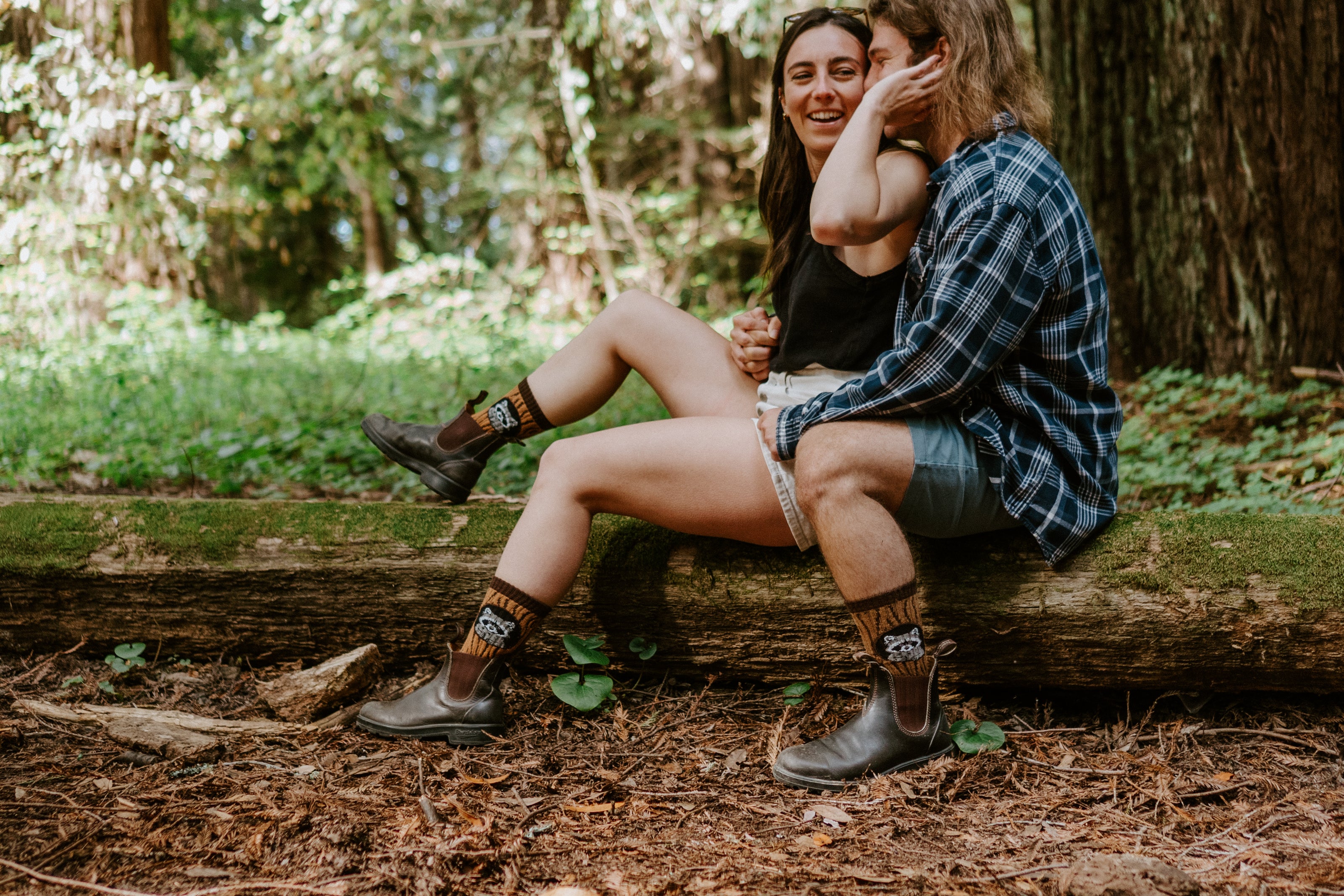 A couple wearing Merino wool raccoon-themed socks sitting closely together on a large fallen log in a forest. The person on the right wears a blue plaid button-up shirt, denim shorts, and dark brown boots, while the person on the left wears a black top, light denim shorts, and patterned socks with dark boots.