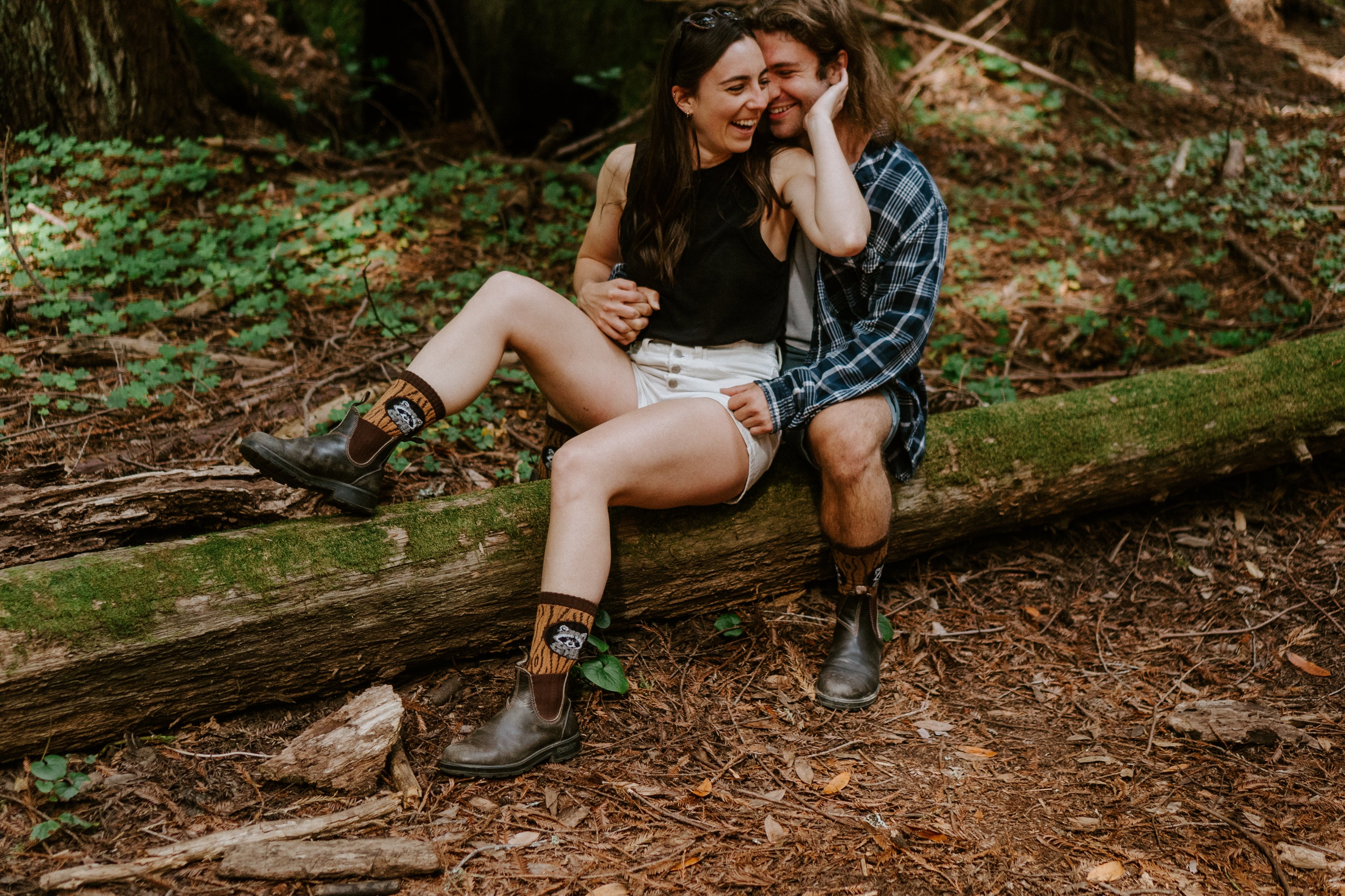 A couple sitting closely together on a large fallen log in a forest, wearing brown leather boots with raccoon-themed patterned socks. 