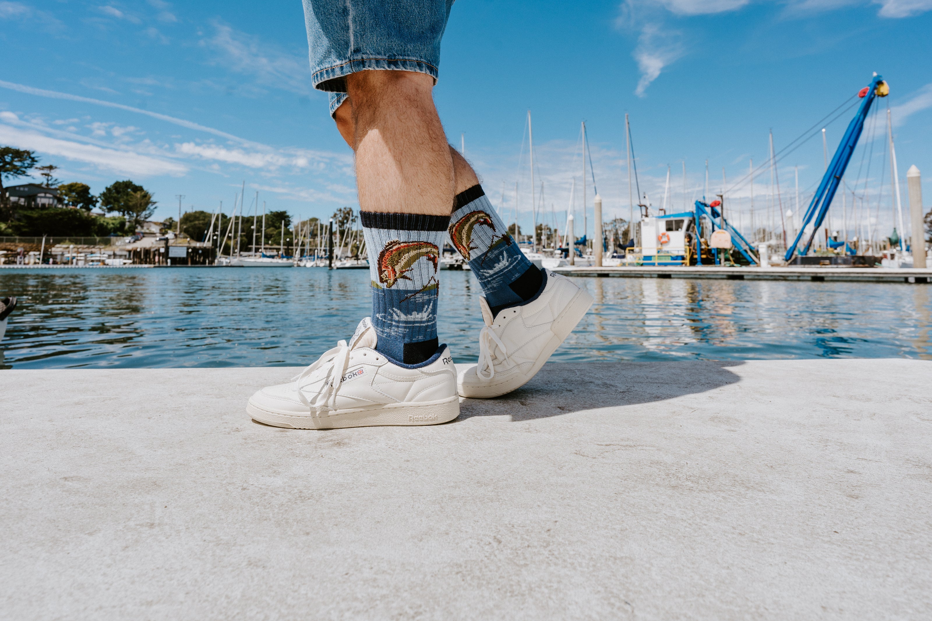 Close-up of a person standing on a concrete dock by a marina, wearing blue jeans, white sneakers, and dark socks with a colorful fish graphic and denim-like texture.
