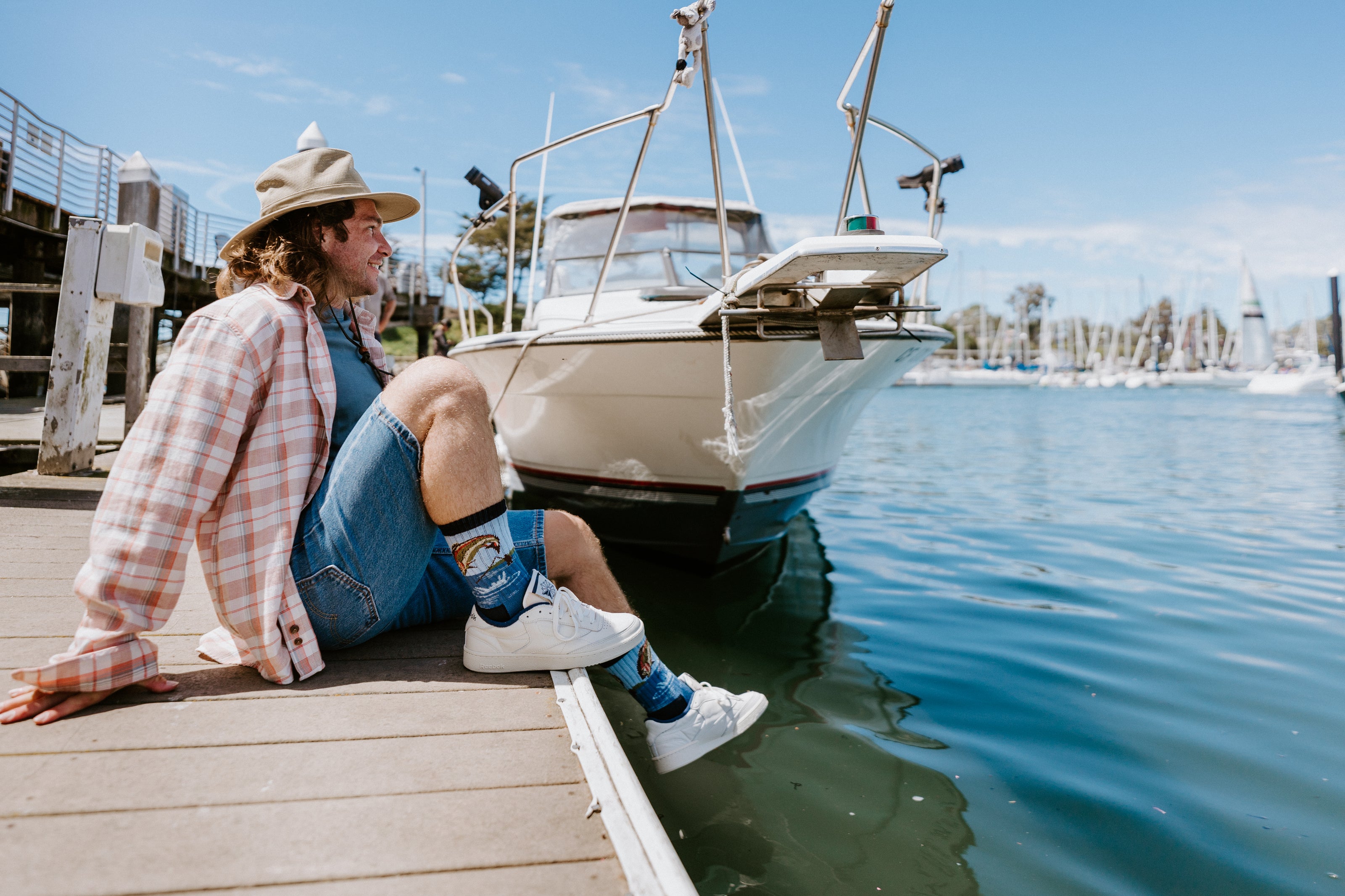 A man wears Merino wool Cast Away socks and sits on a wooden dock next to a boat on calm blue water, wearing a beige hat, a pink and white plaid shirt, denim shorts, and white sneakers.