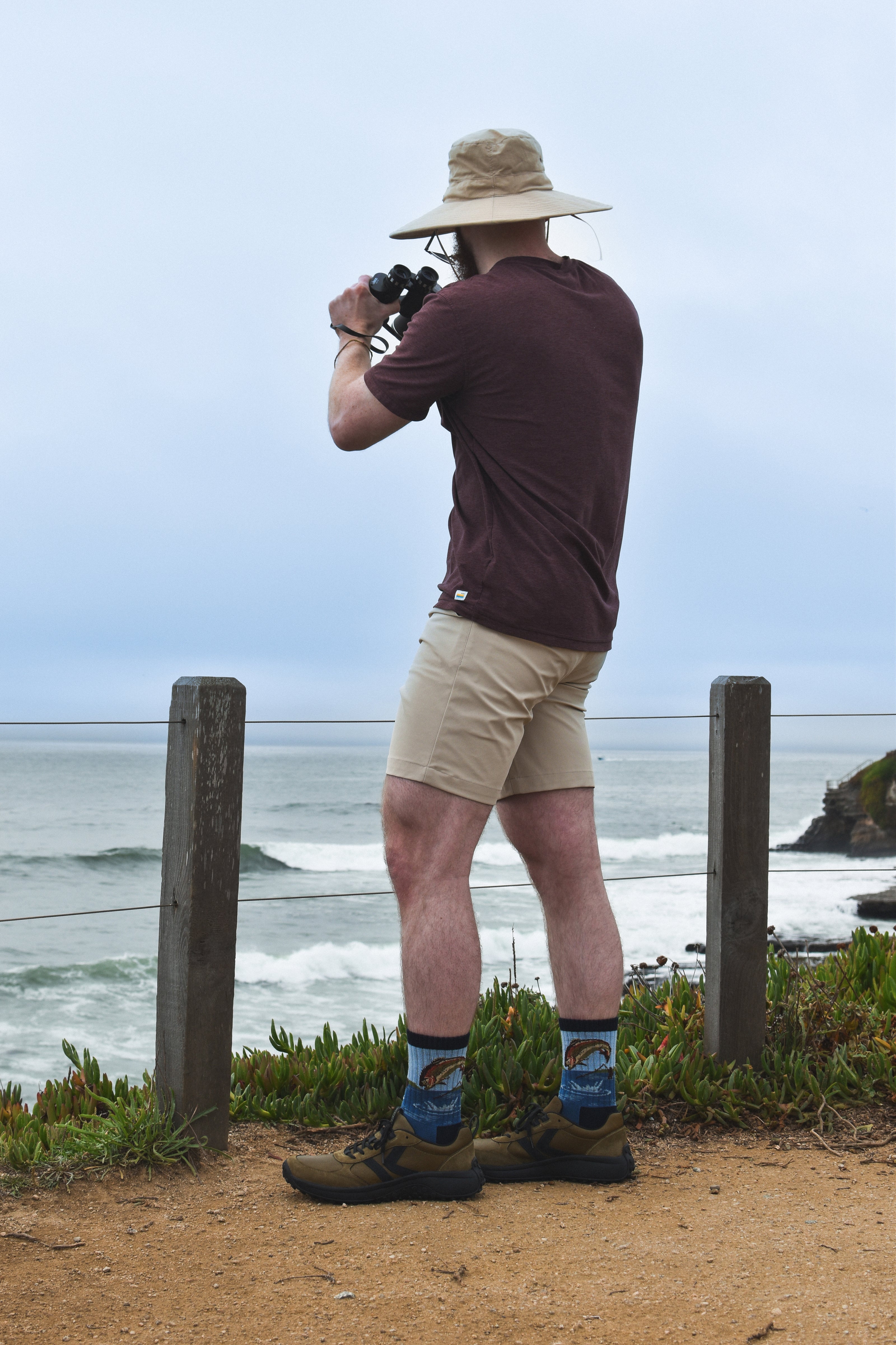 A man wears fish-themed Merino wool socks and stands on a sandy coastal path overlooking the ocean, wearing a beige wide-brim hat, a dark maroon t-shirt, khaki shorts, and hiking boots while holding binoculars.
