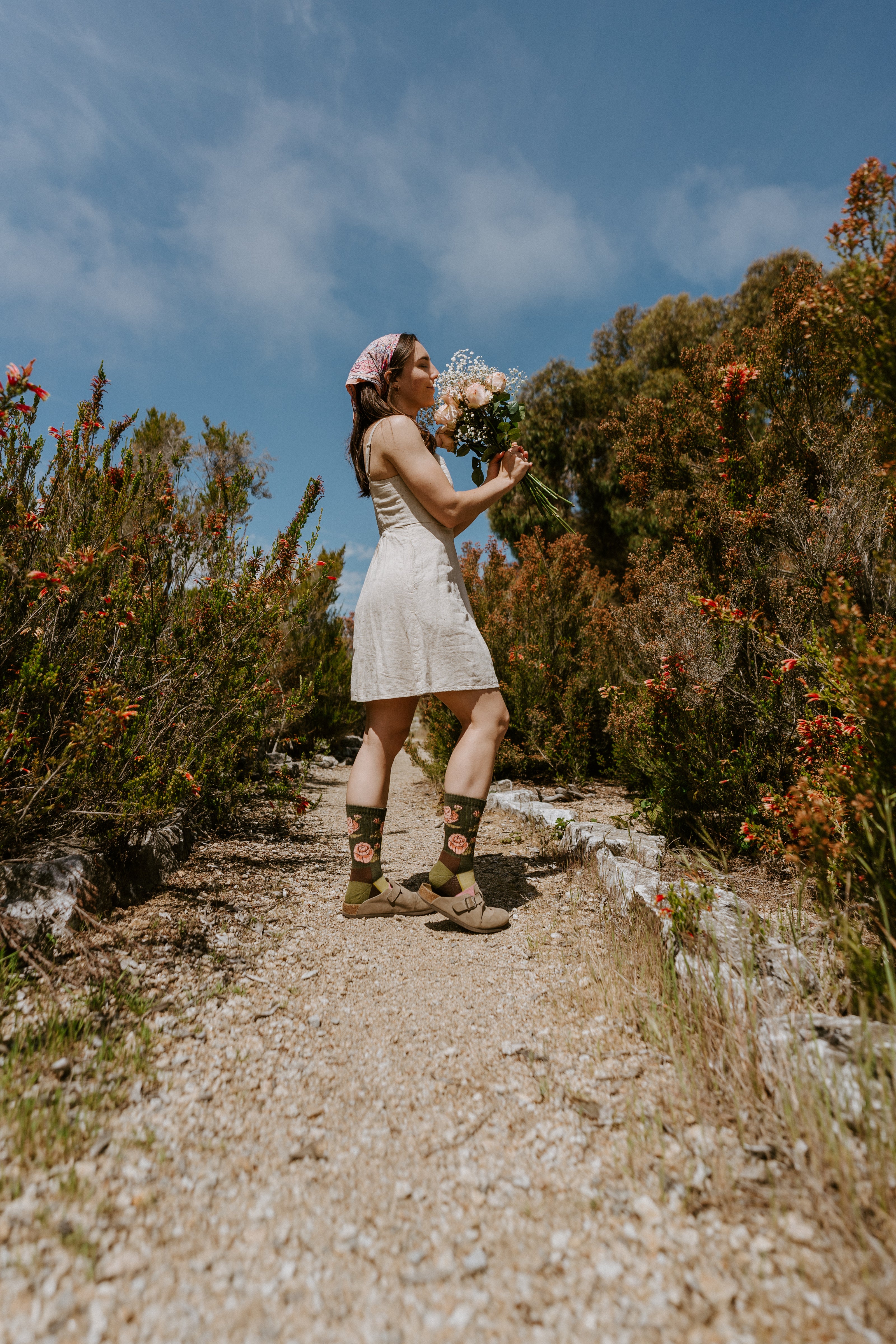 A woman stands on a dirt path outdoors wearing a sleeveless white dress, wearing rose-themed Merino wool socks.