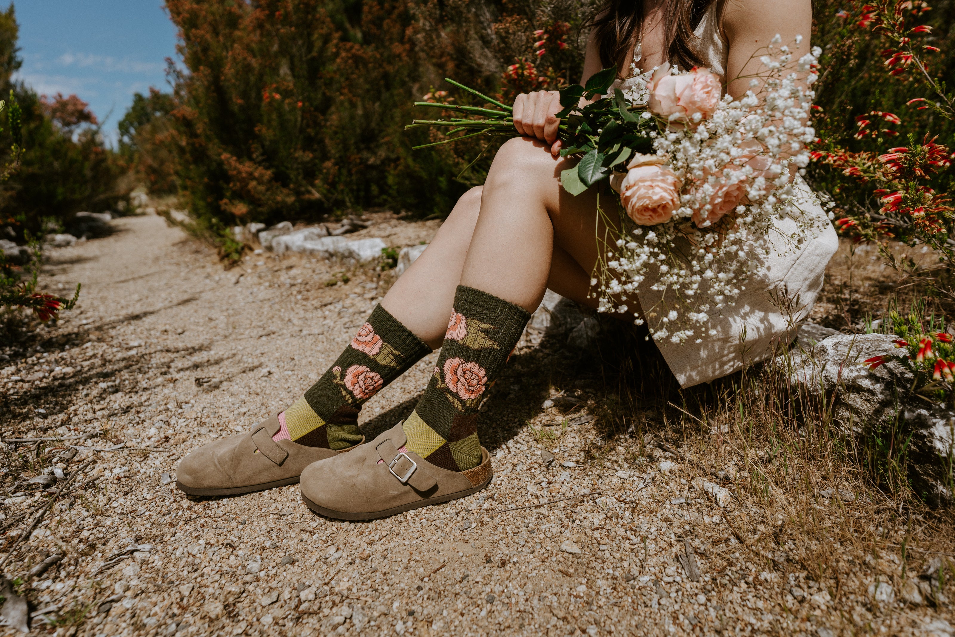 Close-up of a person sitting outdoors on a gravel path wearing dark olive green floral socks with roses on.