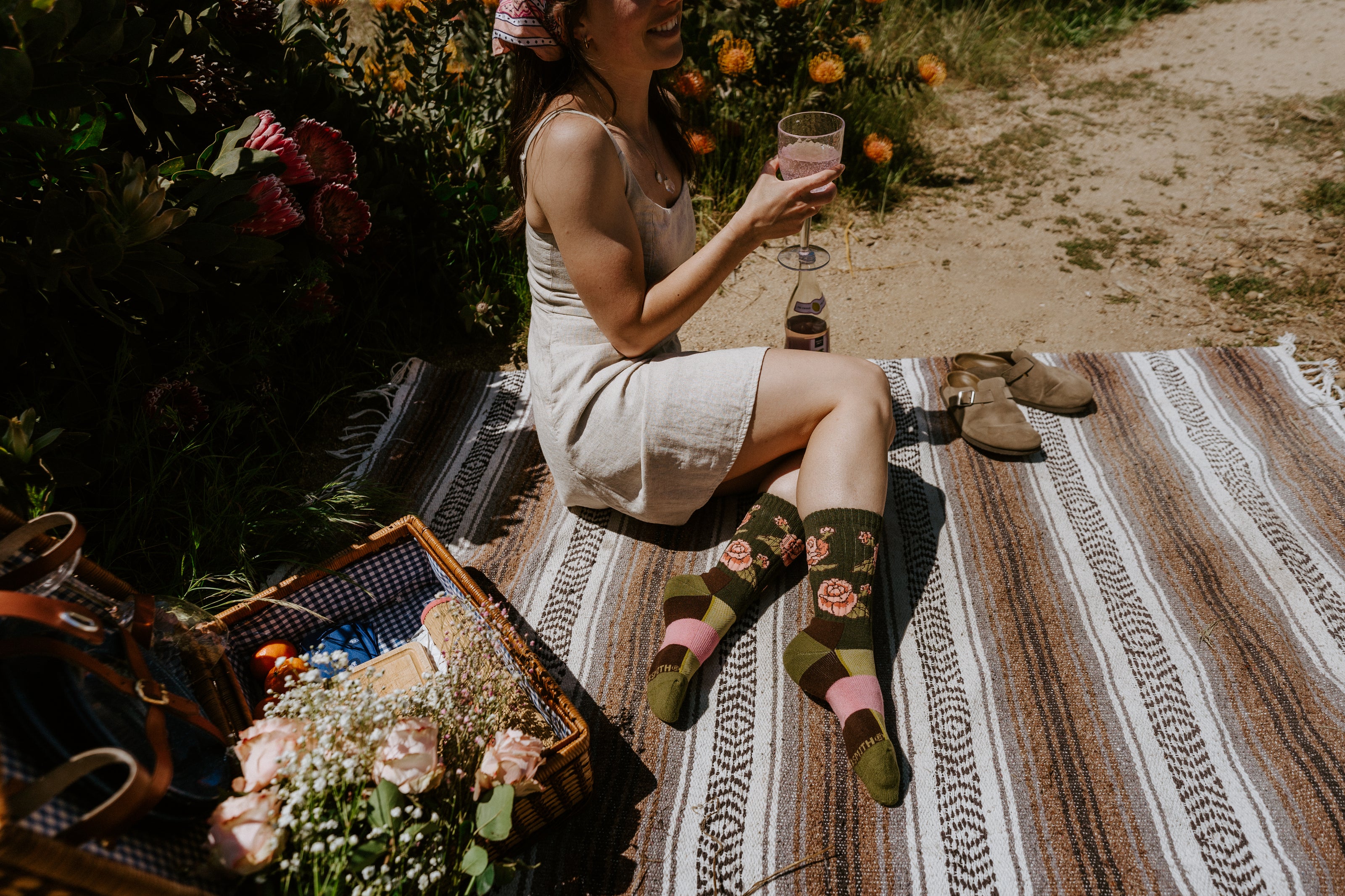 A woman sits on a striped picnic blanket outdoors, wearing dark green rose-themed socks. 