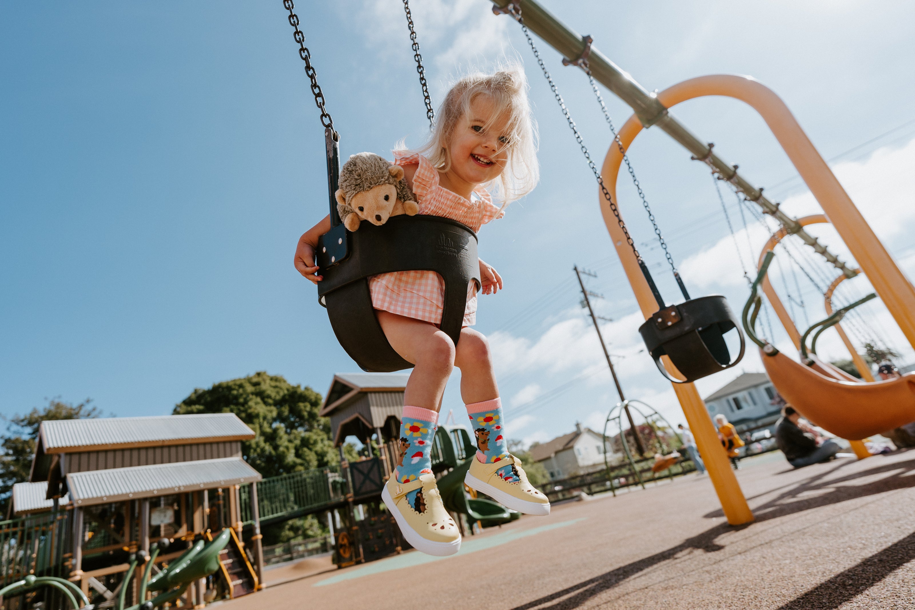 Closeup of child wearing Hedgie Floral socks on a swing set with a stuffed animal and playground equipment in the background