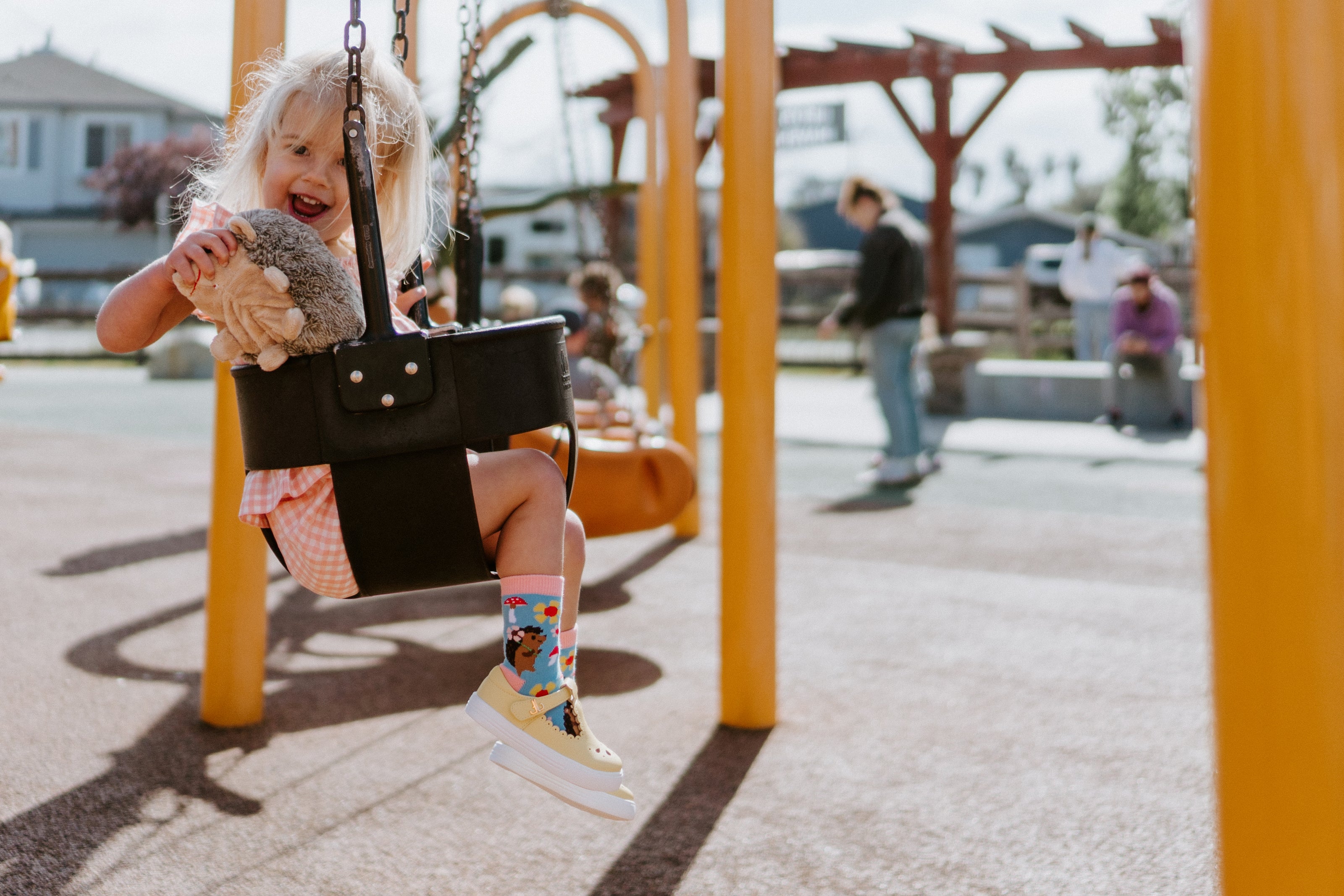 Child wearing Hedgie Floral socks on a swing at a playground holding a teddy bear looking mischievous 