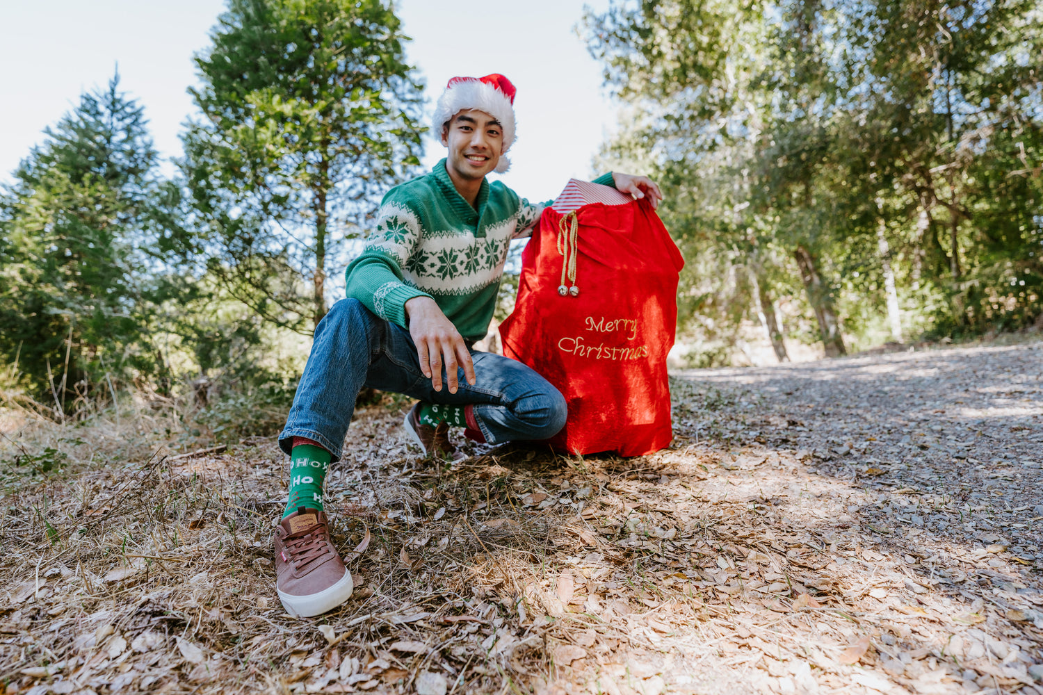 A man wearing a green Christmas sweater, blue jeans, and a Santa hat kneels on a wooded path next to a large red 'Merry Christmas' sack, wearing Ho Ho Ho socks in green