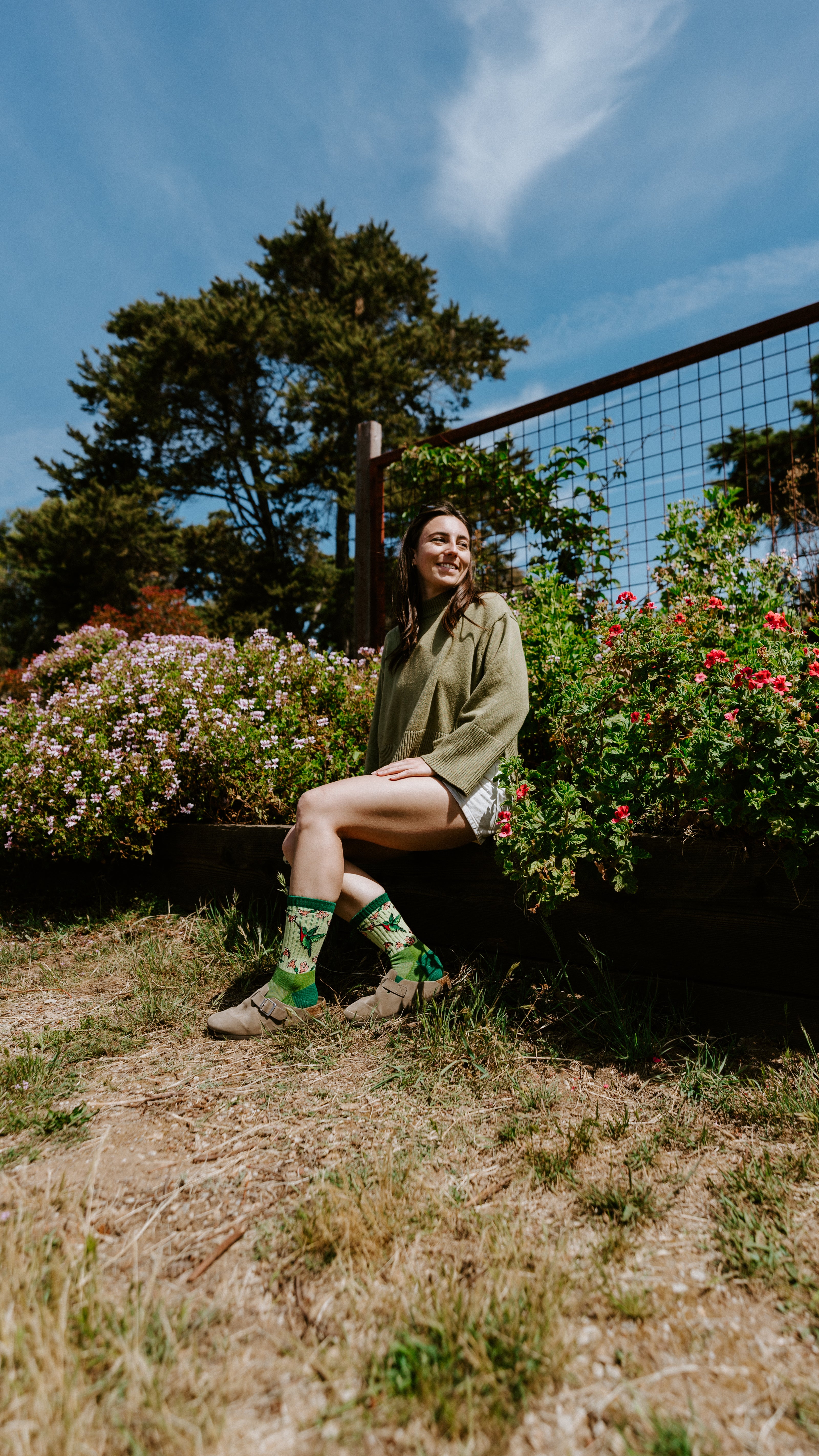 A woman sitting outdoors on a wooden planter box in a garden, wearing hummingbird Merino wool socks.