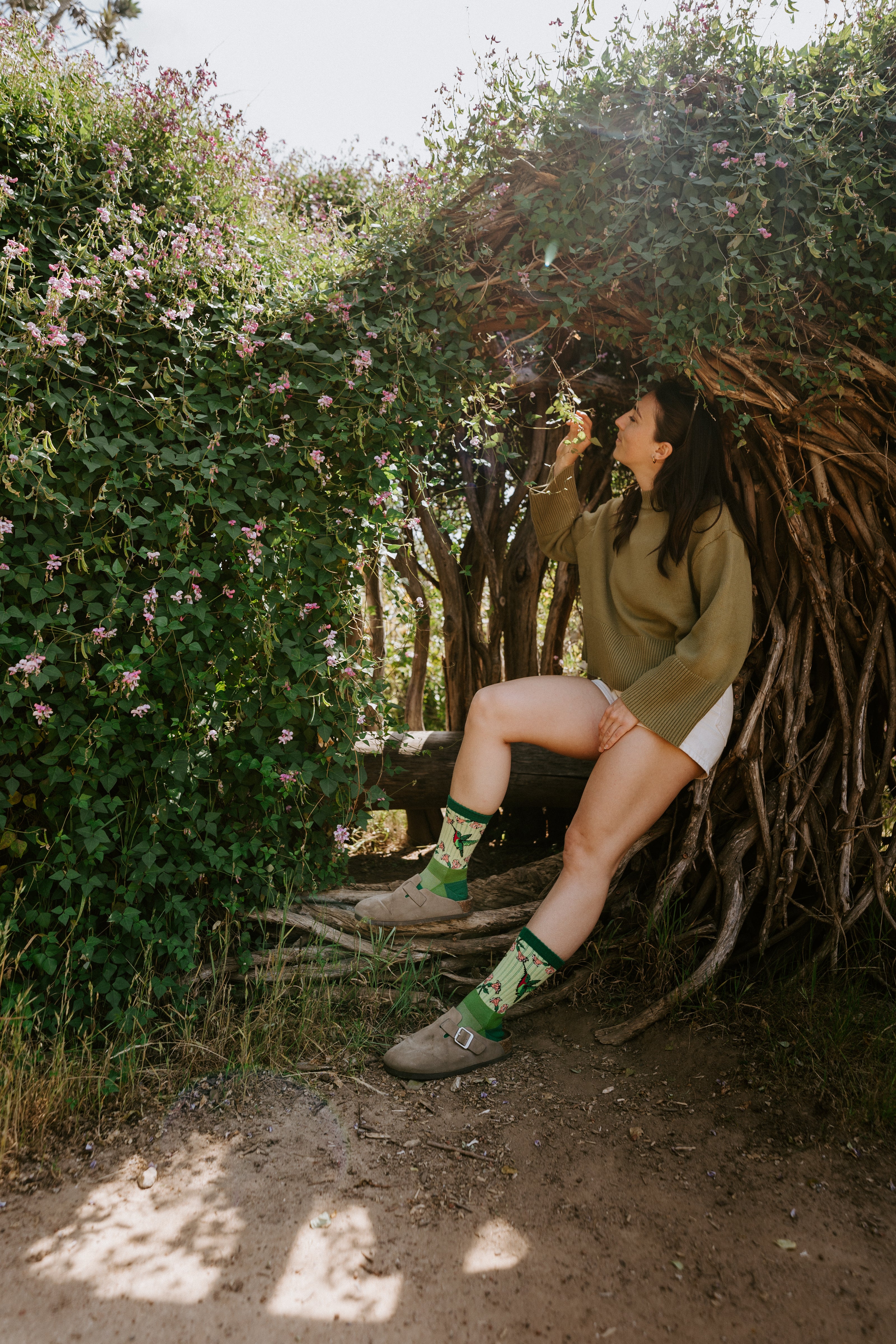 A woman sits on a log in a wooded outdoor setting with dense greenery and pink flowers. She is wearing hummingbird Merino wool socks