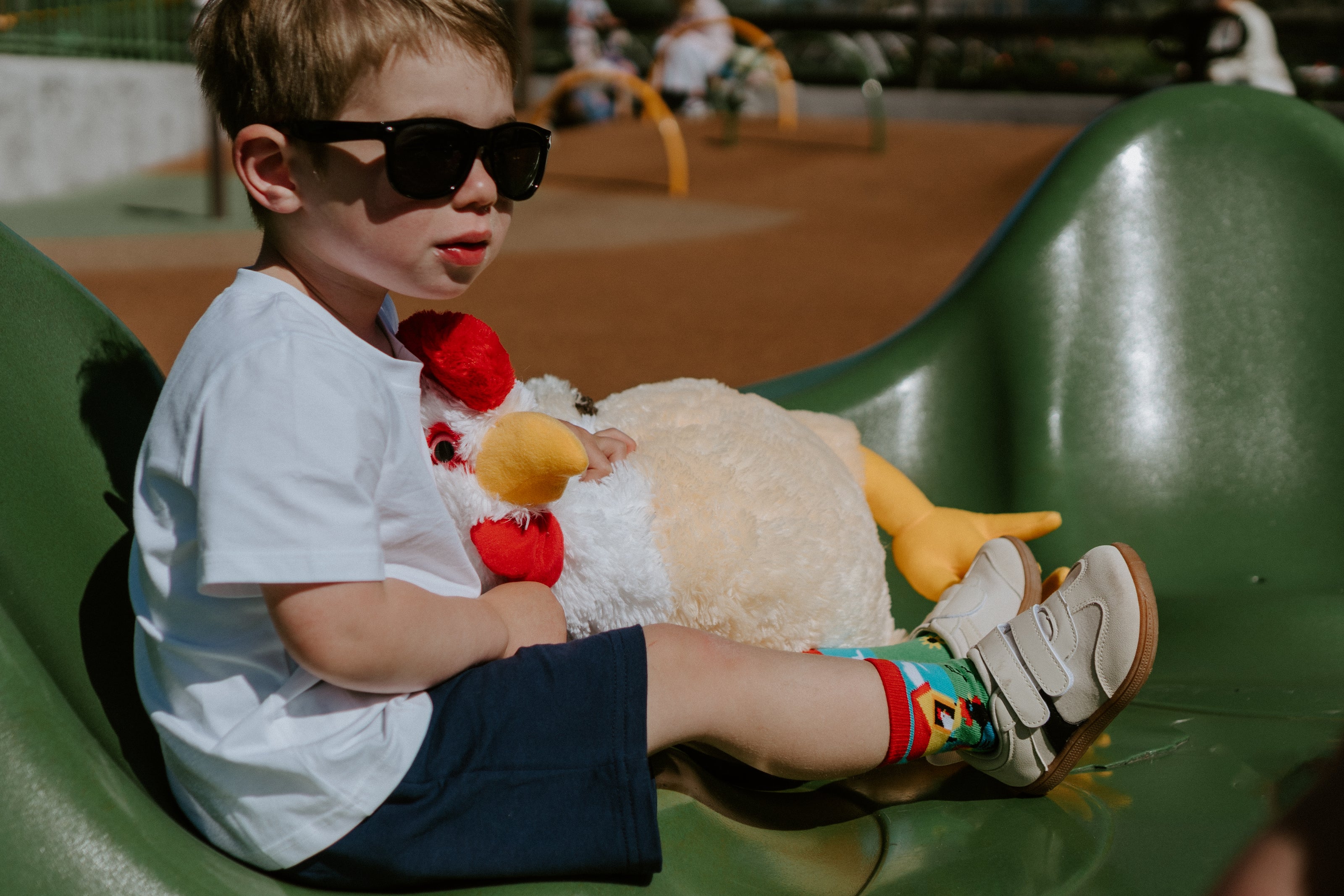 "child wearing sunglasses at playground while holding a chicken stuffed animal and wearing mo chickens kid's socks"