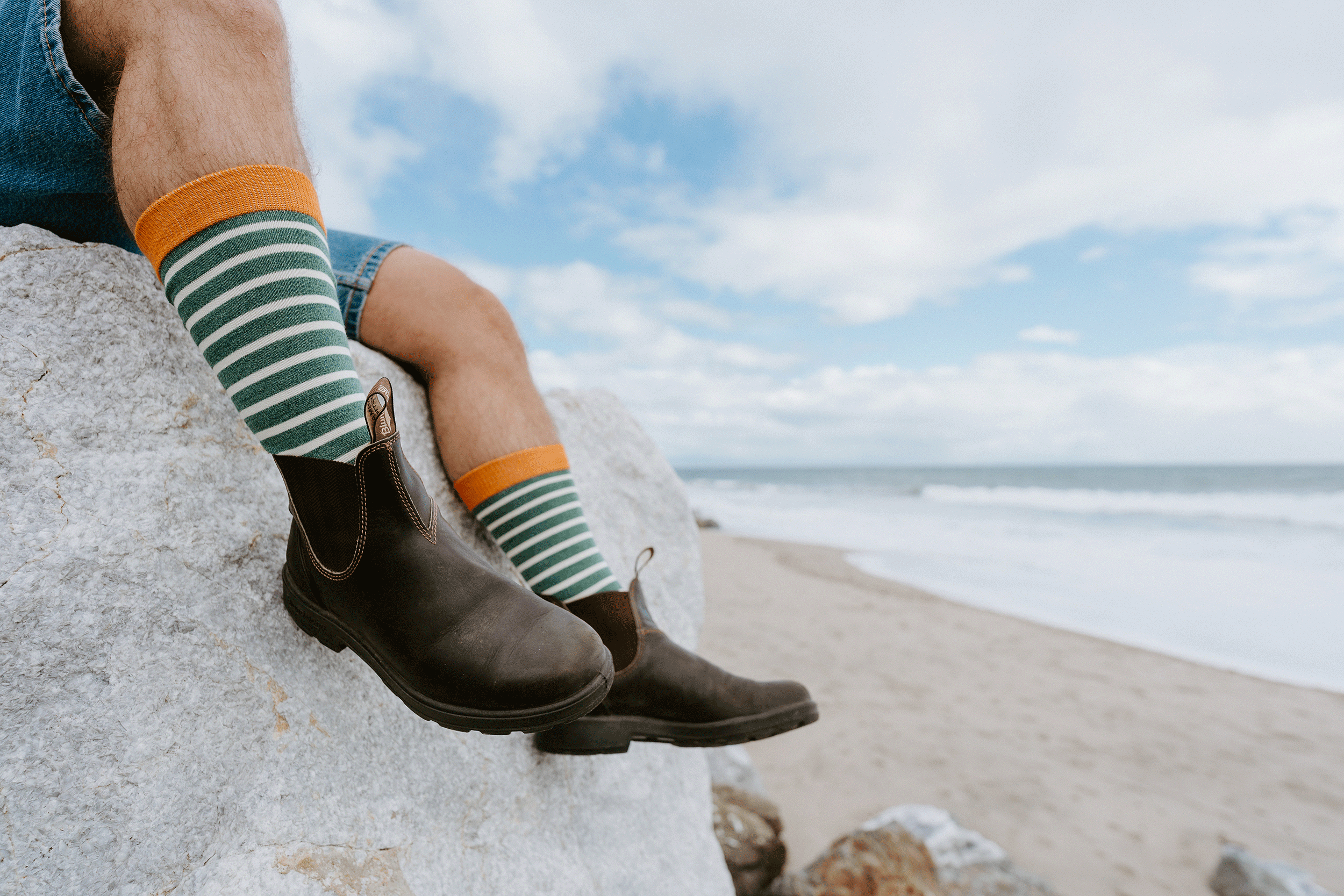 A person sitting on a large rock at the beach, wearing dark brown Chelsea boots, green and white striped socks with orange cuffs, and denim shorts, with the ocean and cloudy sky in the background.