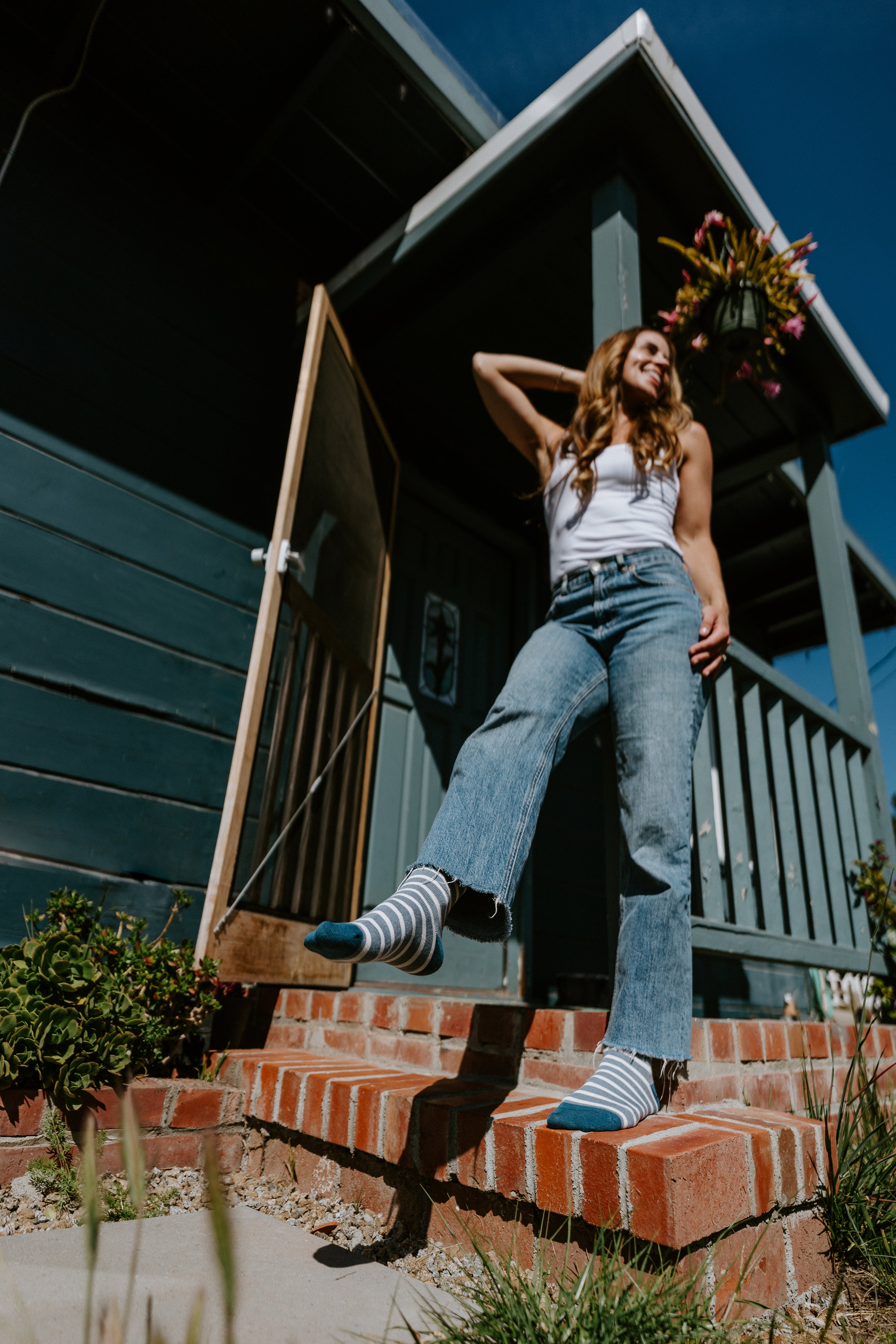 A woman posing on the brick steps of a dark green cabin, wearing blue flared jeans, a white tank top, and blue-and-white bamboo striped socks.
