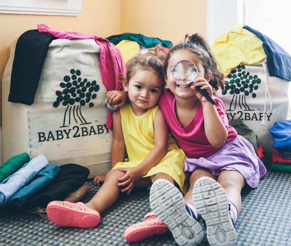 Two children sitting on a floor with a Baby2Baby bag and clothes around them.