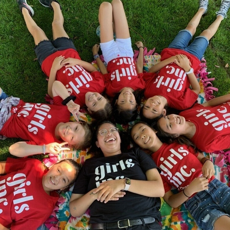 A group of young women wearing "Girls Inc." shirts laying in a circle in the grass with their heads together.
